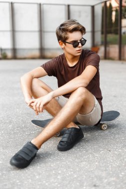 Handsome young man with the hairstyle in fashion sunglasses sitting on a skateboard.