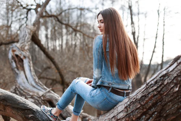 Молодая красивая девушка в джинсовой куртке и джинсах сидит на дереве и мечтает . — стоковое фото