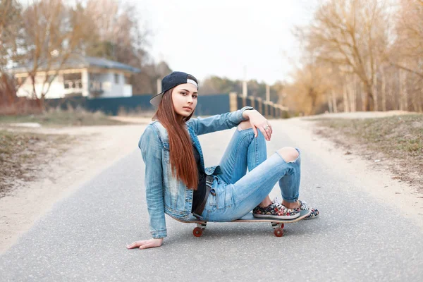 Pretty woman in a baseball cap and denim clothes sitting on a skateboard