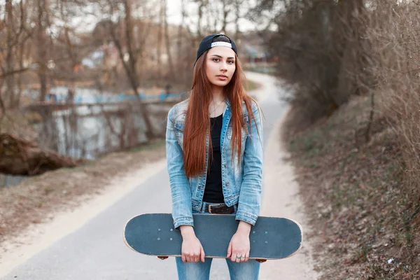 Young beautiful girl in a baseball cap and denim holding a skateboard. on park background — Stock Photo, Image