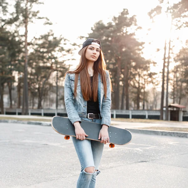 Schöne Frau in Mütze und Jeans mit Skateboard an einem sonnigen Frühlingstag. — Stockfoto