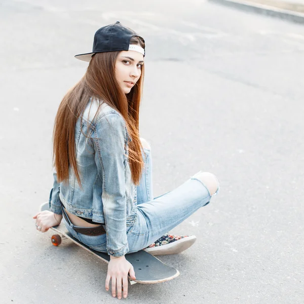 Young stylish girl in a denim jacket and jeans sitting on a skateboard. — Stock Photo, Image