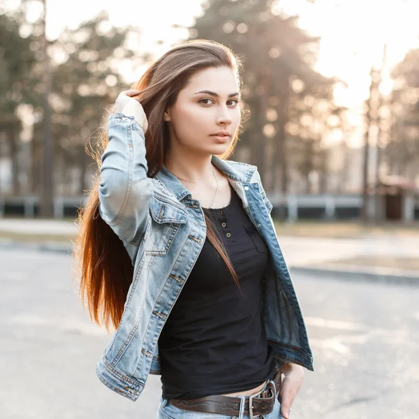 Stylisches junges Mädchen in modischer Jeans-Kleidung korrigiert ihre langen Haare bei Sonnenuntergang — Stockfoto