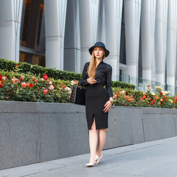 Hermosa mujer elegante en un sombrero elegante en ropa de estilo de negocios sobre un fondo de un edificio moderno —  Fotos de Stock