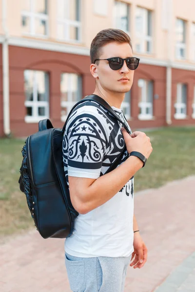 Jovem hipster elegante com uma bolsa e óculos de sol andando pela cidade . — Fotografia de Stock