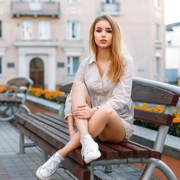 白いスニーカーを履いた小柄な女性が建物の背景のベンチに座っている. — ストック写真