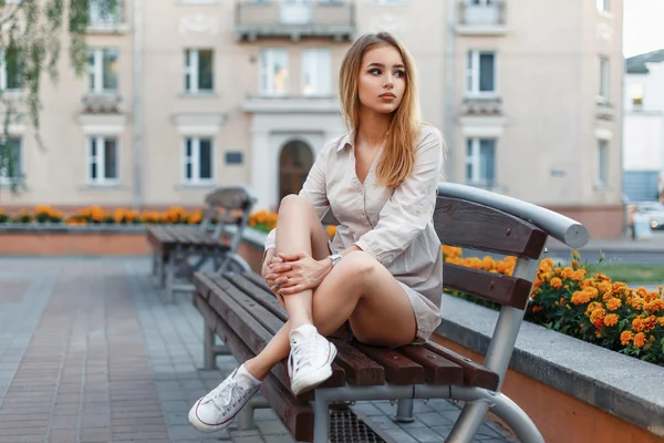 スタイリッシュな服と白いジムの靴の若い美しいファッショナブルな女性がベンチに座って、都市の背景 — ストック写真