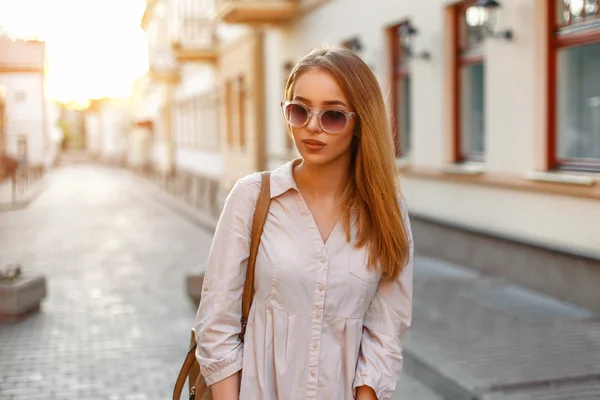 Mulher bonita elegante em roupas da moda e bolsa com óculos de sol em pé ao pôr do sol, fundo urbano — Fotografia de Stock