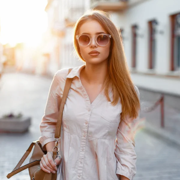 Moda menina elegante em óculos de sol e saco de pé no fundo de um belo pôr do sol . — Fotografia de Stock