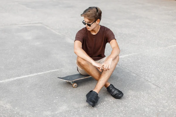 Mladý pohledný muž s účesem ve slunečních brýlích sedí na skateboardu. — Stock fotografie