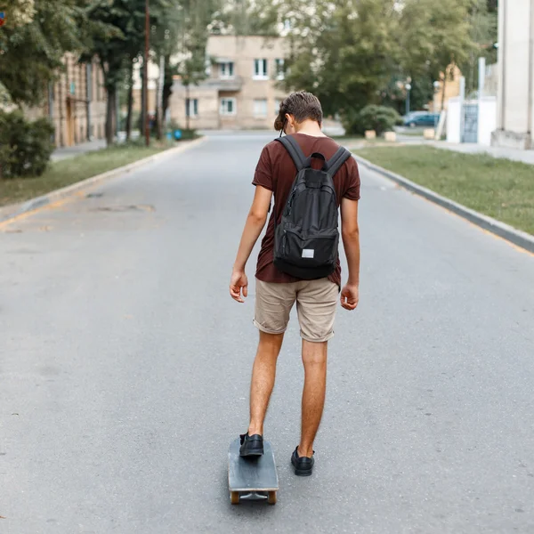 Jeune beau mec avec un sac à dos chevauchant une planche à roulettes sur la route — Photo