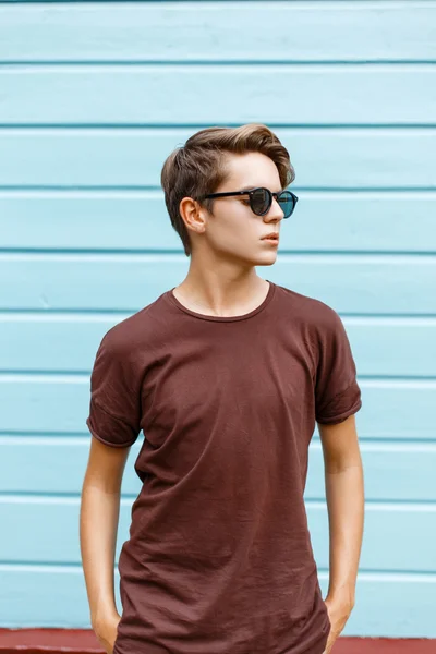 Beau jeune homme avec la coiffure en lunettes de soleil, t-shirt posant près d'un mur en bois bleu vif — Photo