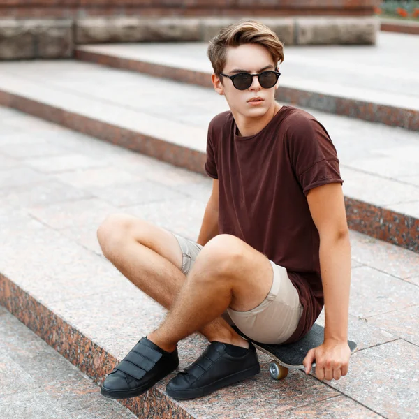 Jeune homme beau en lunettes de soleil reposant sur une planche à roulettes un jour d'été . — Photo