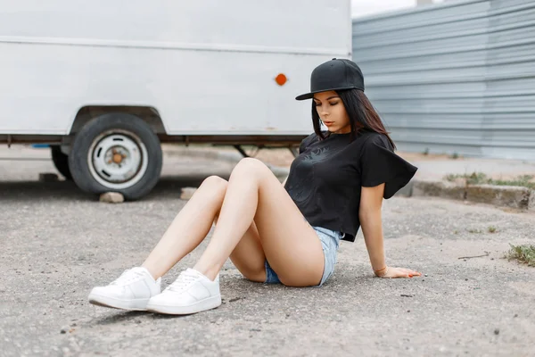 Mooi meisje in een zwarte baseball cap en witte sneakers zitten op de stoep in de buurt van de aanhangwagen. — Stockfoto