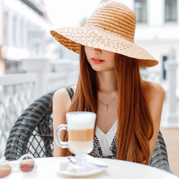 Elegante mujer bonita con labios rojos en un sombrero está sentado en una mesa en un café de verano. En la mesa hay un café y gafas de sol . — Foto de Stock