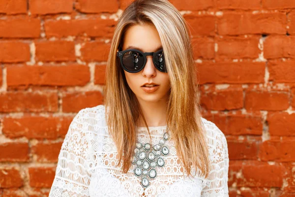 Ritratto di una bella ragazza in camicia di pizzo bianca con occhiali da sole su uno sfondo di un muro di mattoni — Foto Stock