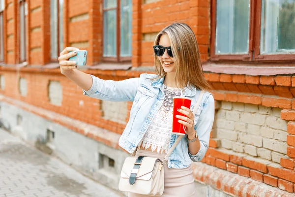 빨간 컵을 들고 있는 데님 재킷을 입고 여름날 전화로 사진을 찍은 아름다운 명랑한 여성. — 스톡 사진