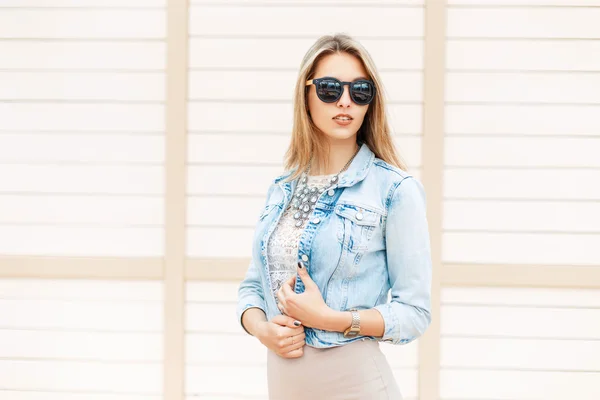 Schöne modische Frau in Sonnenbrille und Jeansjacke, die am Strand in der Nähe der Holzwand steht — Stockfoto