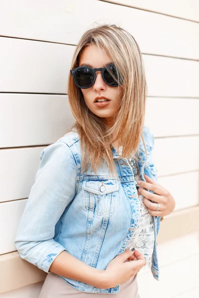 Sexy hermosa chica con estilo en gafas de sol y una chaqueta vaqueros azules de pie cerca de la pared de madera —  Fotos de Stock