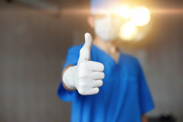 Daumen hoch. Arzt in Maske und blauem Kostüm genehmigt auf einem Hintergrund des Operationssaals. — Stockfoto