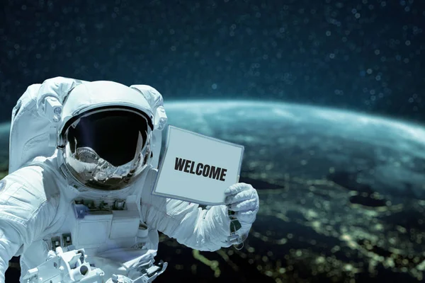 astronaut in a space suit shows a card with the text Welcome spacewalk against the background of the planet Earth. Spaceman in outer space