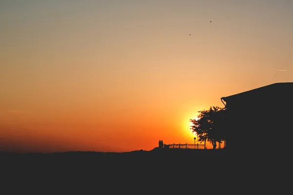 Puesta de sol. Dos personas se paran en una colina con un árbol y una casa Sunse —  Fotos de Stock