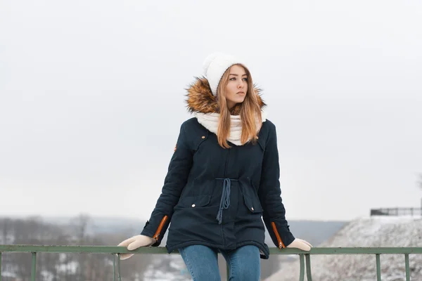 Menina bonito em boné de inverno e casaco sentado no corrimão. Ao ar livre no inverno . — Fotografia de Stock