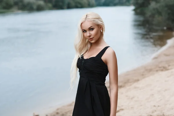 Retrato de una hermosa mujer en un vestido negro en la playa en el río — Foto de Stock