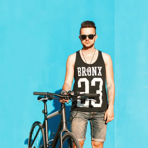 Jovem bonitão em óculos de sol, uma camiseta preta e shorts jeans montando uma bicicleta. No fundo de uma parede azul brilhante . — Fotografia de Stock