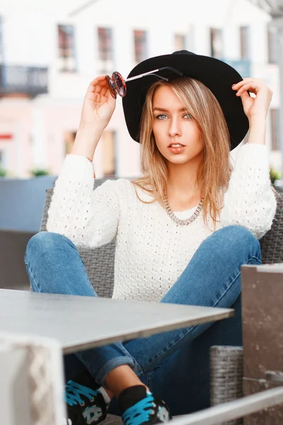 Retrato de uma jovem mulher bonita com um chapéu preto e óculos de sol redondos. Menina bonita senta-se no café de verão . — Fotografia de Stock