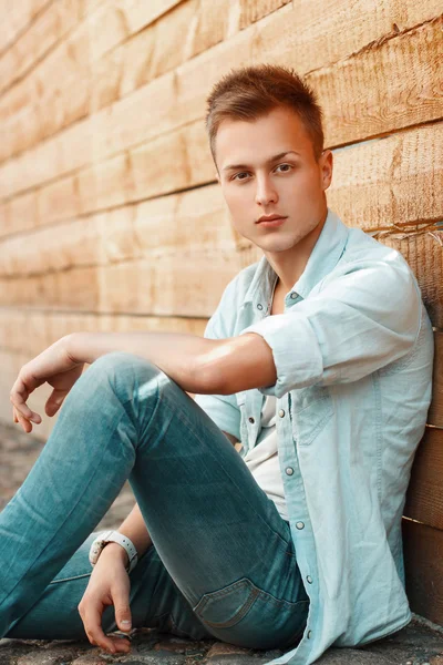 Jovem homem bonito em jeans roupas sentadas perto de uma parede de madeira . — Fotografia de Stock