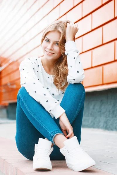 Портрет молодой красивой девушки в белом свитере, джинсах и белых кроссовках, сидящей на плитке возле оранжевой стены — стоковое фото