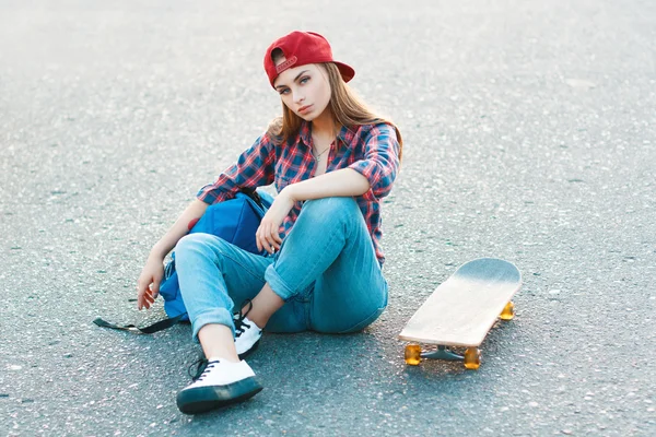 Beautiful young and fashionable woman with a bag and skateboard sitting on the pavement. — 图库照片