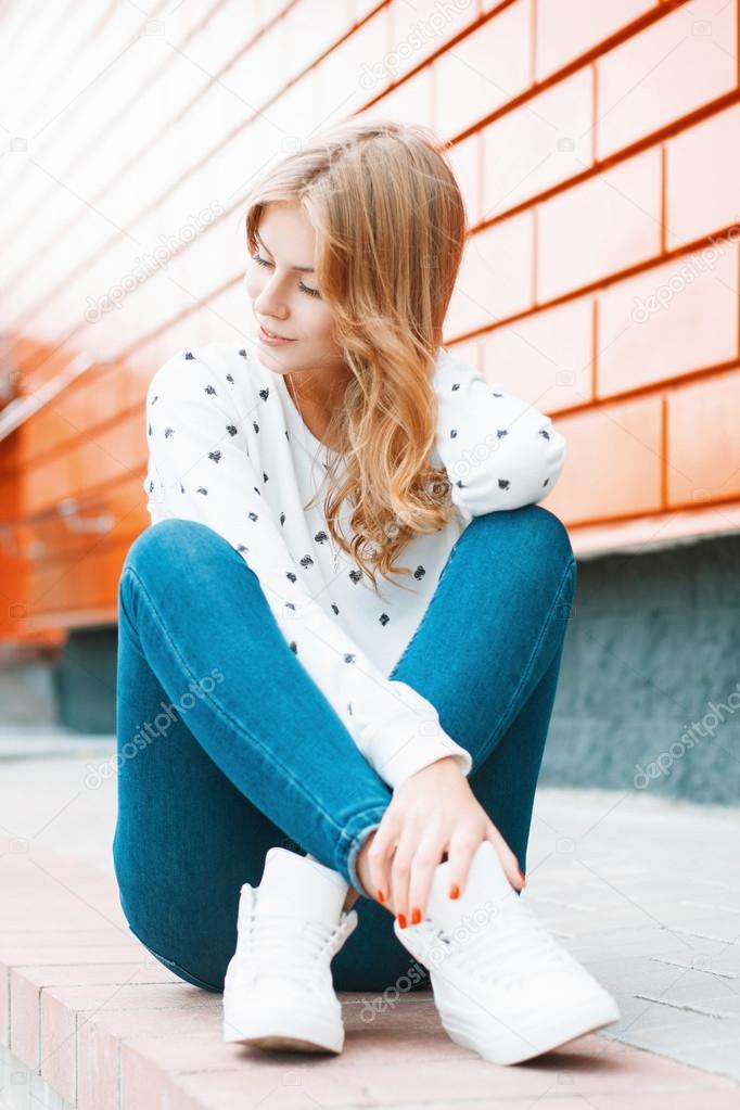 Beautiful woman in a white sweater, jeans and white sneakers sitting on a tile near the orange wall