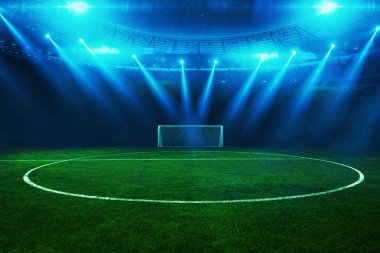 Geceleri, boş yeşil alan ve kale direğinin üzerinde parlayan spot ışıklarıyla aydınlık bir futbol stadyumu dramatik ve enerjik bir atmosfer yaratıyor. Futbol turnuvası ve futbol şampiyonları