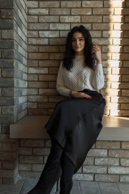 Doğal ışıkta tuğla bir duvarın yanında oturan zarif kadın beyaz bir kazak ve siyah etek giyiyor. Havalı bir sonbahar moda portresi. İçerde sakin ve kendine güvenen bir ifade.