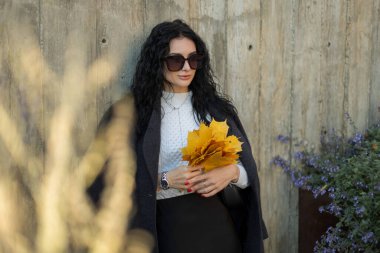 Sonbahar demeti sarı yapraklı güneş gözlüklü, şık bir ceket ve süveter giyen, dışarıda poz veren genç bir kadın. Sonbahar havası, zarafet ve modern sokak tarzı..