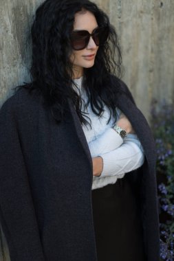 Uzun kıvırcık saçlı, gri ceketli, dışarıda, güneş ışığında duran güzel bir esmer. Çağdaş moda ve yaşam tarzı fotoğrafçılığı.
