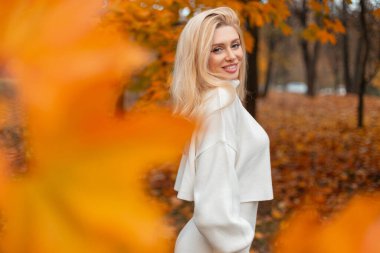 Sonbahar parkında beyaz kazaklı, gülen sarışın kadın altın yapraklarla dışarıda poz veriyor, sonbahar mevsiminin ve ılık doğal atmosferin tadını çıkarıyor.