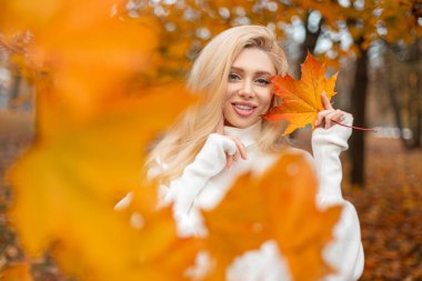 Sonbahar yeşilliklerinde açık havada poz veren beyaz kazaklı genç bir kadın. Pozitif trend kızı parlak sarı bir akçaağaç yaprağı tutuyor. Sonbahar havası ve havası.