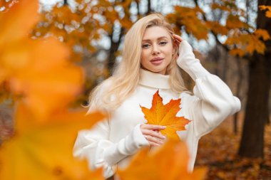 Sarı akçaağaç yaprağıyla sonbahar mevsiminde dışarıda poz veren güzel sarışın. Pozitif trend kızı parlak sarı bir akçaağaç yaprağı tutuyor. Sonbahar havası ve havası.