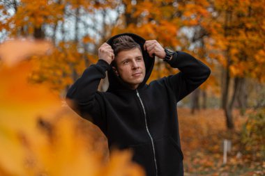 Sonbahar parkında turuncu yapraklarla çevrili siyah kapüşonlu genç adam yürüyor. Havalı siyah kapüşonlu bir adam bir sonbahar parkında yürürken kapüşonu takıyor..