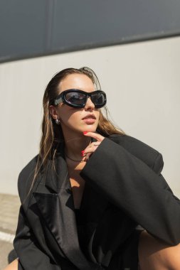Güneş ışınları altında açık havada poz veren siyah ceketli göz kamaştırıcı güneş gözlüklü güzel şehirli genç kadın modeli. Modaya uygun bir bayan.