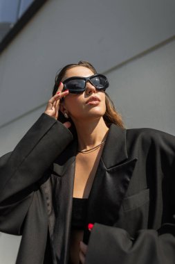 Büyük beden ceketli ve aksesuarlı modaya uygun giyinmiş genç bir bayan model, gün ışığında dışarıda poz veriyor. Siyah elbiseli güzel kız şehirde güneş gözlüğü takıyor.