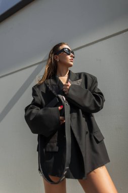 Güneş gözlüklü ve büyük beden ceketli moda bir kadın bina duvarının yanında duruyor. Gün ışığında minimalist moda portresi.