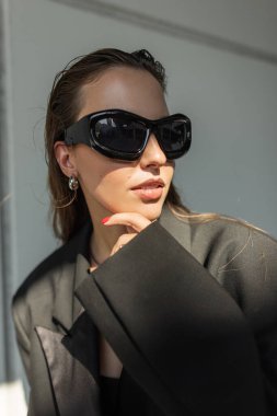 Büyük boy siyah ceketli ve güneş gözlüklü genç bir kadın açık havada poz veriyor, modern sokak tarzı moda portresi kendine güvenen ve modaya uygun görünümlü..