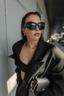 Aşırı büyük siyah ceket ve kalın güneş gözlüğü takan modacı kadın model, açık havada minimalist şehir geçmişiyle poz veriyor. Modaya uygun siyah çanta.