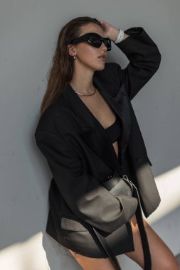 Şık, büyük beden ceketli, modaya uygun güneş gözlüklü, kentin arka planına karşı özgüvenle poz veren şık bir kadın modeli. Modaya uygun siyah çanta.