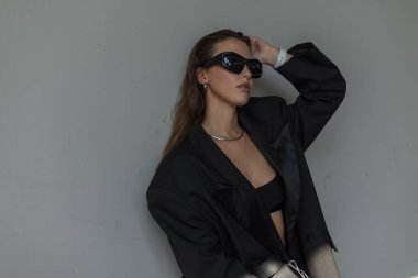 Büyük beden ceketli, fütüristik güneş gözlüklü şık bir kadının portresi, doğal güneş ışığında minimalist şehir tarzı..