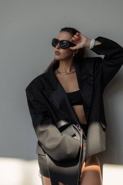 Büyük beden ceketli, şık güneş gözlüklü ve zarif makyajlı zarif genç bir manken. Güneş ışığında şık poz veriyor..